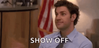 A Man In A Suit And Tie Is Sitting In Front Of An American Flag And Says `` Show Off '' . GIF