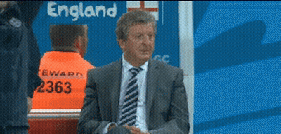 A Man In A Suit And Tie Is Sitting In Front Of An England Sign GIF