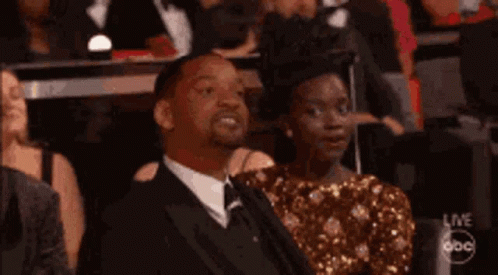 A Man In A Suit And Tie Is Sitting Next To A Woman In A Gold Dress In A Crowd . GIF