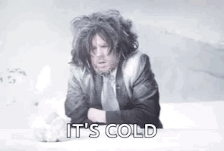 A Man In A Suit And Tie Is Sitting On A Bed With His Arms Crossed And Says `` It 'S Cold '' . GIF