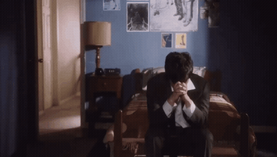 A Man In A Suit And Tie Is Sitting On A Bed With His Hands Folded . GIF