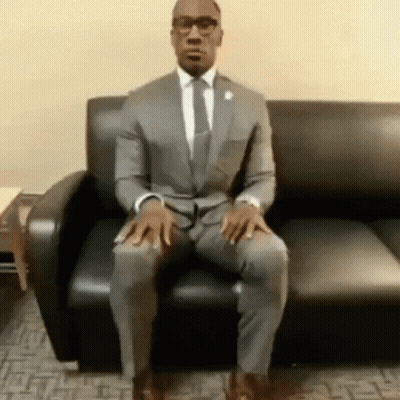 A Man In A Suit And Tie Is Sitting On A Black Couch . GIF