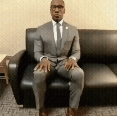 A Man In A Suit And Tie Is Sitting On A Black Couch With His Legs Crossed . GIF