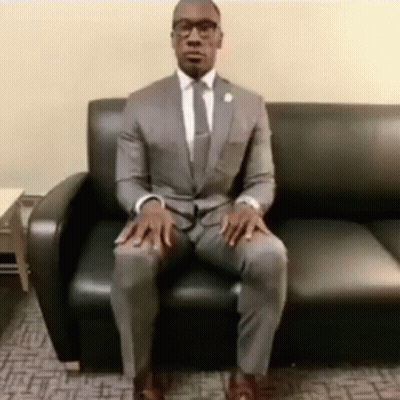 A Man In A Suit And Tie Is Sitting On A Black Couch With His Legs Crossed . GIF