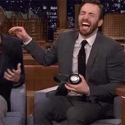 A Man In A Suit And Tie Is Sitting On A Couch Laughing And Holding A Camera . GIF