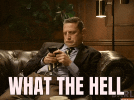 A Man In A Suit And Tie Is Sitting On A Couch Looking At His Cell Phone And The Words What The Hell Are Above Him GIF