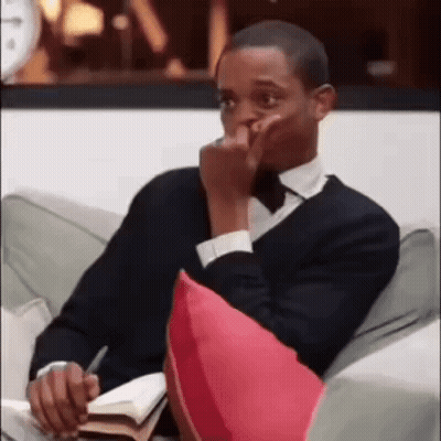 A Man In A Suit And Tie Is Sitting On A Couch With A Book And A Pen In His Hand . GIF