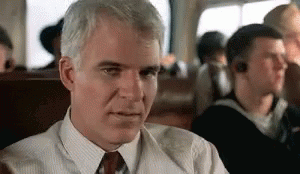 A Man In A Suit And Tie Is Sitting On A Plane Looking At Something . GIF
