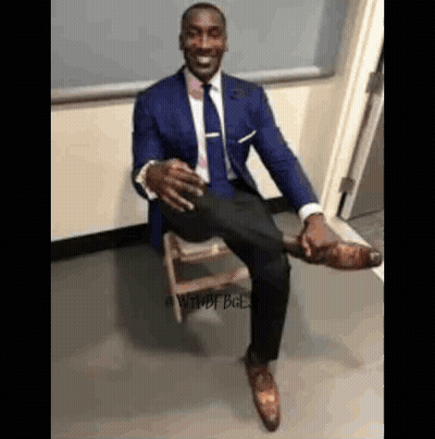 A Man In A Suit And Tie Is Sitting On A Wooden Stool With His Leg Up . GIF