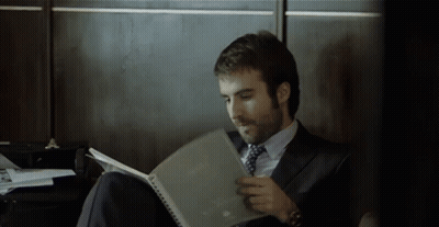 A Man In A Suit And Tie Is Sitting On The Floor Reading A Book GIF