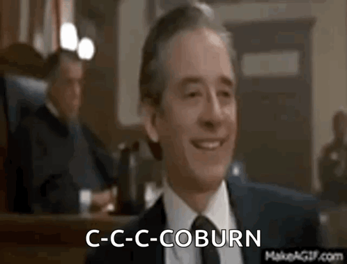 A Man In A Suit And Tie Is Smiling In A Courtroom While Another Man Sits Behind Him . GIF
