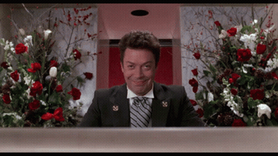 A Man In A Suit And Tie Is Smiling In Front Of A Display Of Flowers GIF