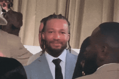 A Man In A Suit And Tie Is Smiling In Front Of A Group Of People GIF