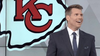 A Man In A Suit And Tie Is Smiling In Front Of A Large Kc Logo GIF
