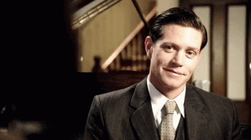 A Man In A Suit And Tie Is Smiling In Front Of A Piano . GIF