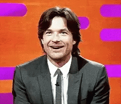 A Man In A Suit And Tie Is Smiling In Front Of A Purple And Orange Background . GIF