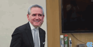 A Man In A Suit And Tie Is Smiling In Front Of A Tv And A Can Of G12 GIF