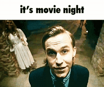 A Man In A Suit And Tie Is Smiling In Front Of A Woman In A White Dress And The Words It 'S Movie Night Below Him GIF
