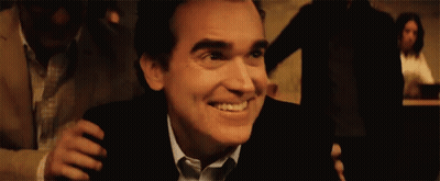 A Man In A Suit And Tie Is Smiling While Being Hugged By Another Man In A Dark Room . GIF