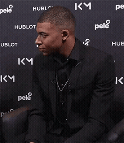 A Man In A Suit And Tie Is Smiling While Sitting In Front Of A Wall With Logos On It . GIF