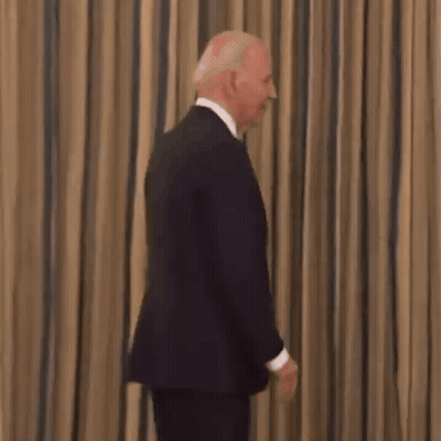 A Man In A Suit And Tie Is Smiling While Standing In Front Of A Striped Curtain . GIF