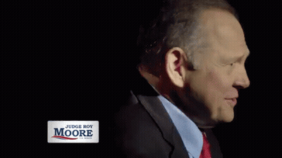 A Man In A Suit And Tie Is Smiling With A Judge Roy Moore Sign Behind Him GIF