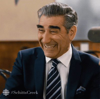 A Man In A Suit And Tie Is Smiling With #Schitts Creek In The Upper Right Corner GIF