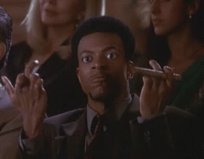 A Man In A Suit And Tie Is Smoking A Cigar In Front Of A Crowd . GIF