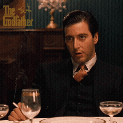 A Man In A Suit And Tie Is Smoking A Cigarette While Sitting At A Table In Front Of A Poster For The Godfather GIF