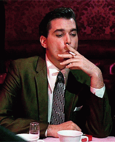 A Man In A Suit And Tie Is Smoking A Cigarette While Sitting At A Table GIF