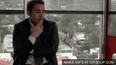 A Man In A Suit And Tie Is Smoking A Cigarette While Sitting In Front Of A Window . GIF