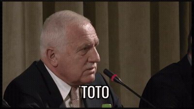A Man In A Suit And Tie Is Speaking Into A Microphone And The Word Toto Is On The Screen Behind Him GIF