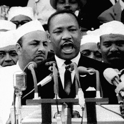 A Man In A Suit And Tie Is Speaking Into A Microphone In Front Of A Crowd Of People . GIF