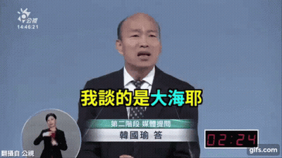 A Man In A Suit And Tie Is Speaking Into A Microphone While A Woman In Sign Language Watches GIF