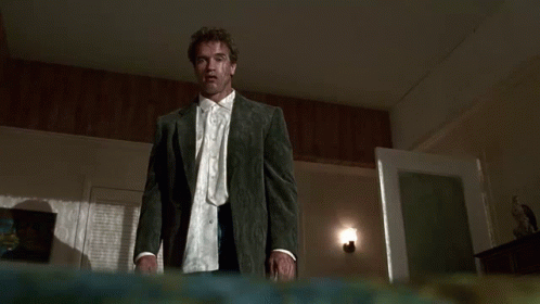 A Man In A Suit And Tie Is Standing In A Dark Room And Looking At The Camera . GIF