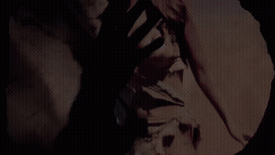 A Man In A Suit And Tie Is Standing In A Dark Room Looking Out Of A Hole In The Wall . GIF