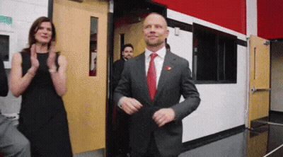 A Man In A Suit And Tie Is Standing In A Doorway With A Woman Clapping Behind Him GIF