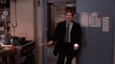 A Man In A Suit And Tie Is Standing In A Kitchen Holding A Cup And A Flashlight . GIF