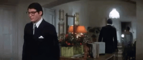 A Man In A Suit And Tie Is Standing In A Room Next To A Table . GIF