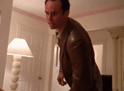 A Man In A Suit And Tie Is Standing In A Room With A Lamp And A Door . GIF