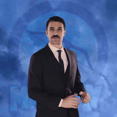 A Man In A Suit And Tie Is Standing In Front Of A Blue Background With The Letter M. GIF