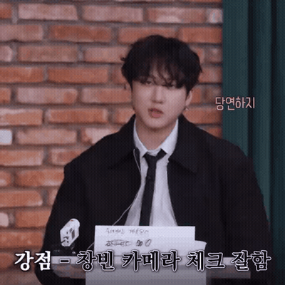 A Man In A Suit And Tie Is Standing In Front Of A Brick Wall And Holding A Piece Of Paper With Korean Writing On It GIF