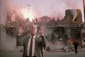 A Man In A Suit And Tie Is Standing In Front Of A Building That Is On Fire . GIF