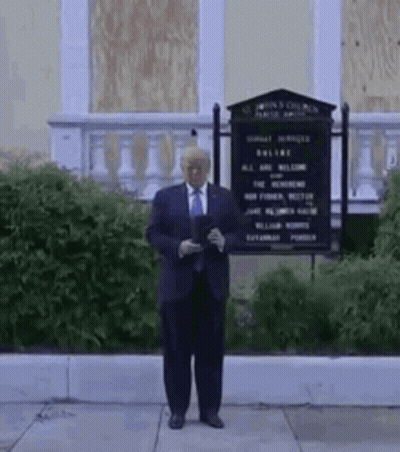 A Man In A Suit And Tie Is Standing In Front Of A Building With A Sign . GIF