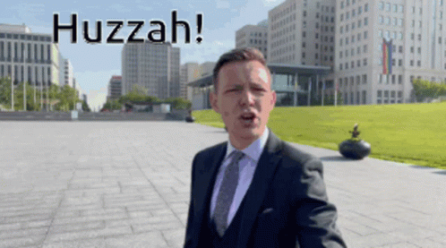 A Man In A Suit And Tie Is Standing In Front Of A City And The Words Huzzah Are Above Him GIF
