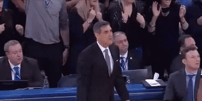 A Man In A Suit And Tie Is Standing In Front Of A Crowd Of People At A Basketball Game . GIF