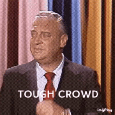 A Man In A Suit And Tie Is Standing In Front Of A Curtain And Says Tough Crowd . GIF