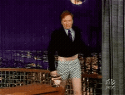 A Man In A Suit And Tie Is Standing In Front Of A Curtain That Says Nbc GIF
