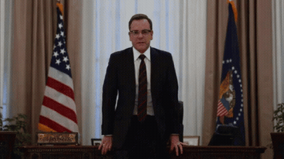 A Man In A Suit And Tie Is Standing In Front Of A Desk In A Room With Flags . GIF