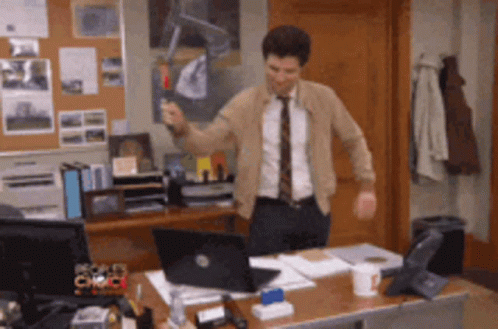 A Man In A Suit And Tie Is Standing In Front Of A Desk With A Laptop GIF